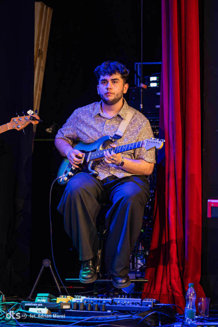 Musician seated on stage playing a blue electric guitar, with a pedalboard at his feet and red stage curtains beside him.