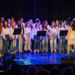 Chorus of students wearing white tops and jeans performing on stage as a conductor leads them from the front (concert setting).