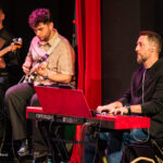 Three musicians performing on a stage: bassist left, guitarist center, pianist with red Nord keyboard on the right; banner in back.