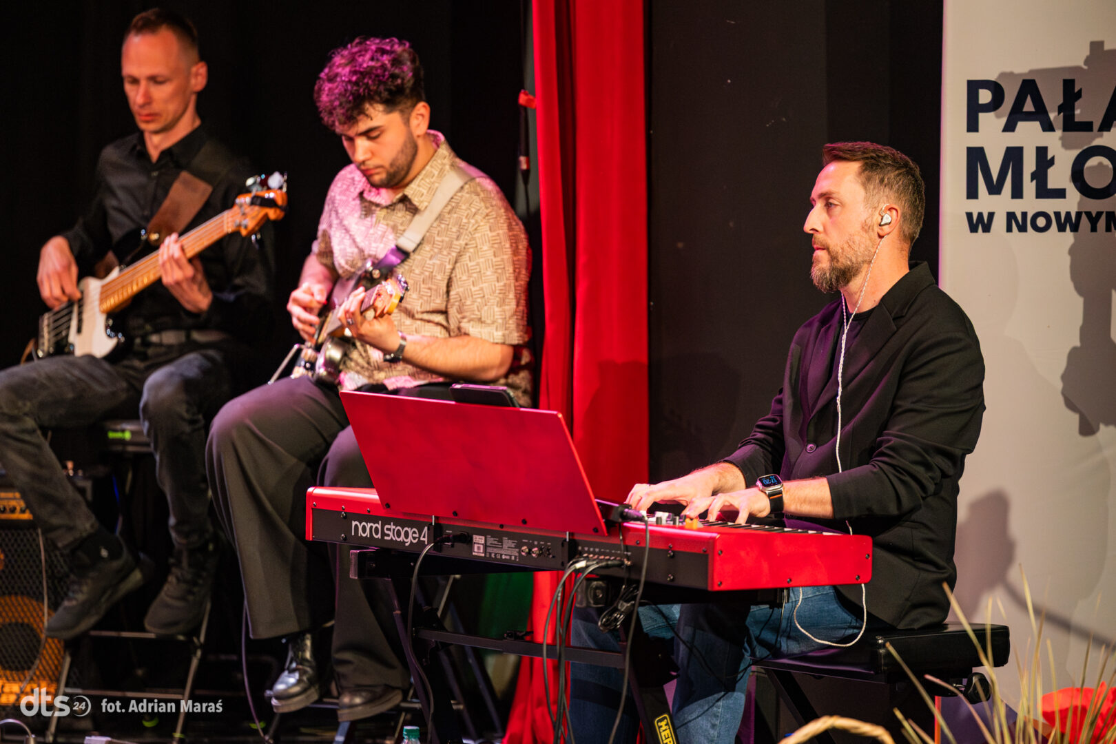 Three musicians performing on a stage: bassist left, guitarist center, pianist with red Nord keyboard on the right; banner in back.