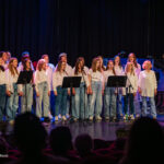 Children's choir on stage singing, led by a female conductor in a white lace dress on the right.