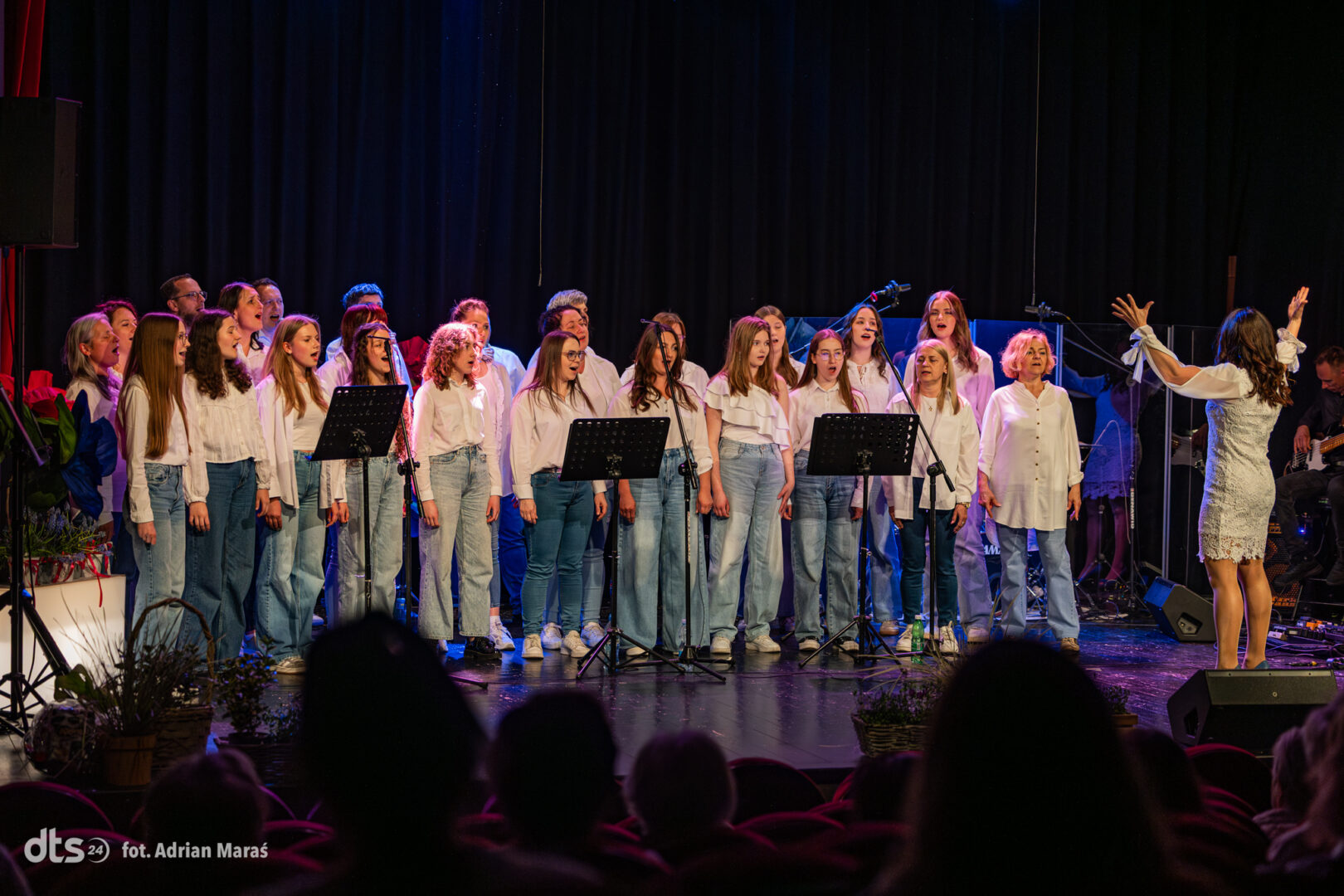 Children's choir on stage singing, led by a female conductor in a white lace dress on the right.
