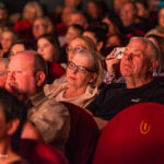 Audience seated in a dim cinema, watching a performance or film together in red upholstered seats.