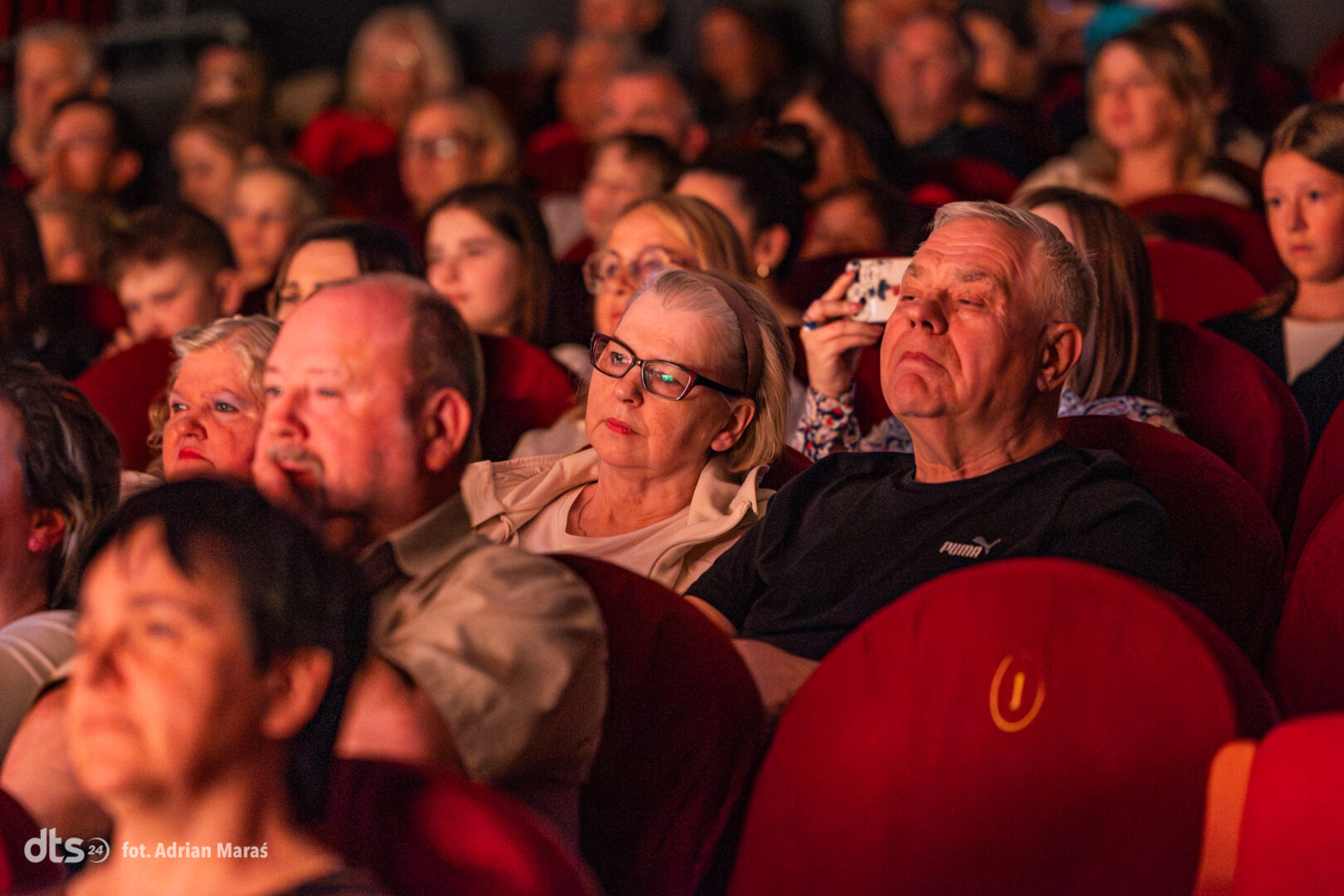 Audience seated in a dim cinema, watching a performance or film together in red upholstered seats.