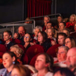 Audience seated in red theater seats, watching a performance with warm stage lighting on their faces.