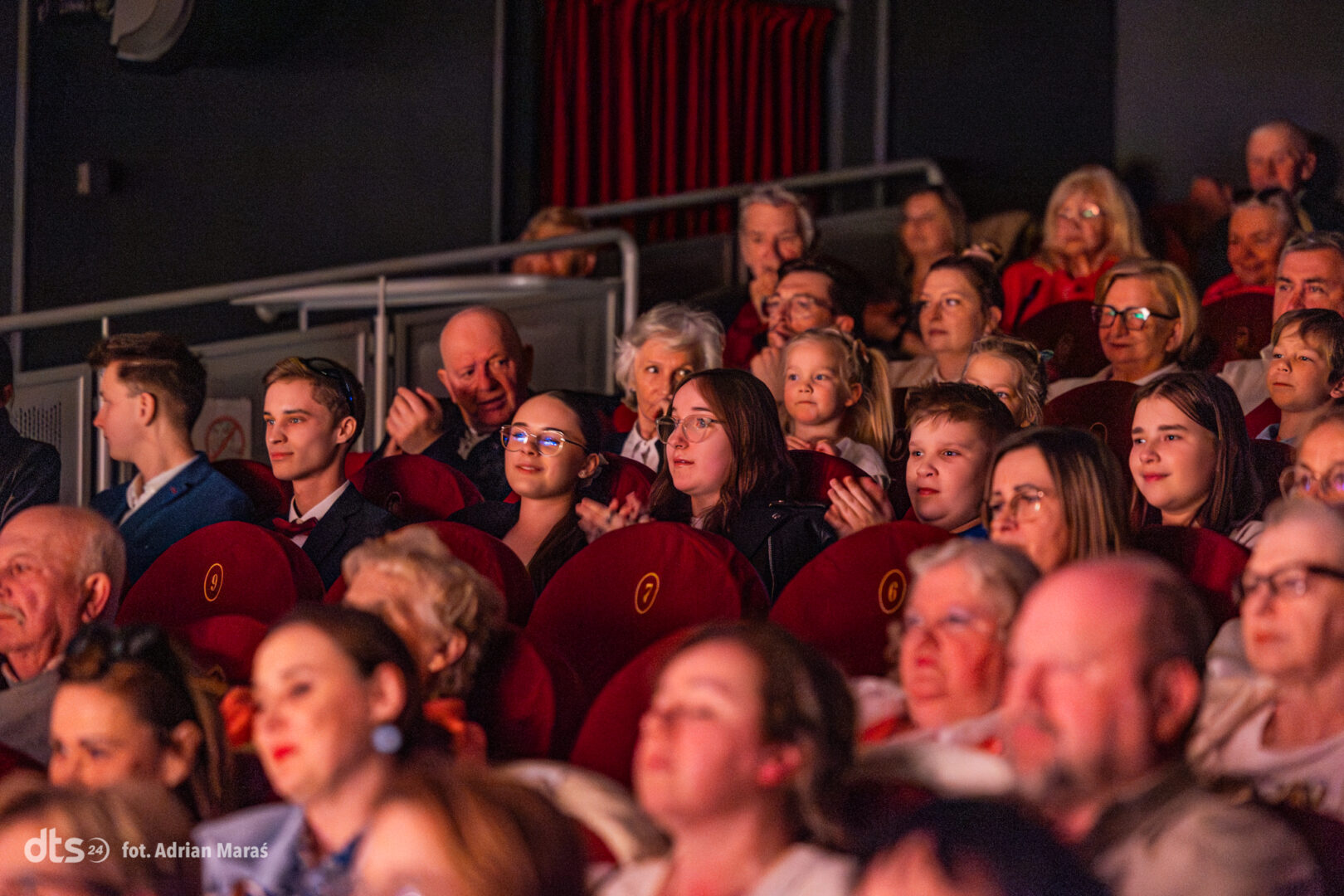 Audience seated in red theater seats, watching a performance with warm stage lighting on their faces.