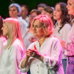 A chorus on stage, front and center an older woman with short blond hair claps as fellow singers in white shirts look on under pink stage lights.