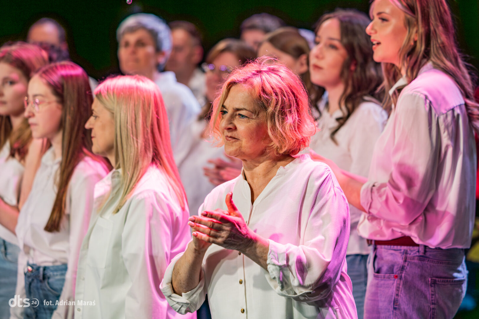 A chorus on stage, front and center an older woman with short blond hair claps as fellow singers in white shirts look on under pink stage lights.