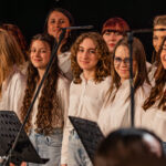 Group of teenage girls in white shirts and jeans singing on stage, with microphone stands in front.