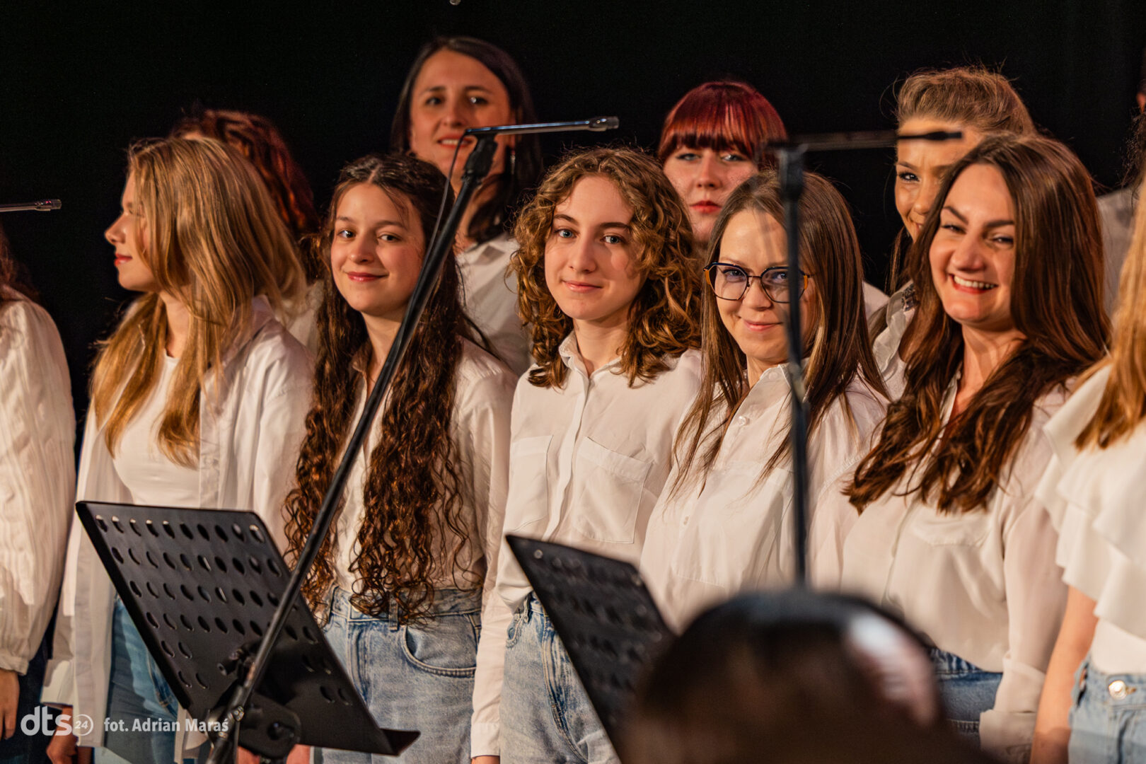 Group of teenage girls in white shirts and jeans singing on stage, with microphone stands in front.