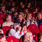 Audience in a theater, applauding and watching a performance from red seats.