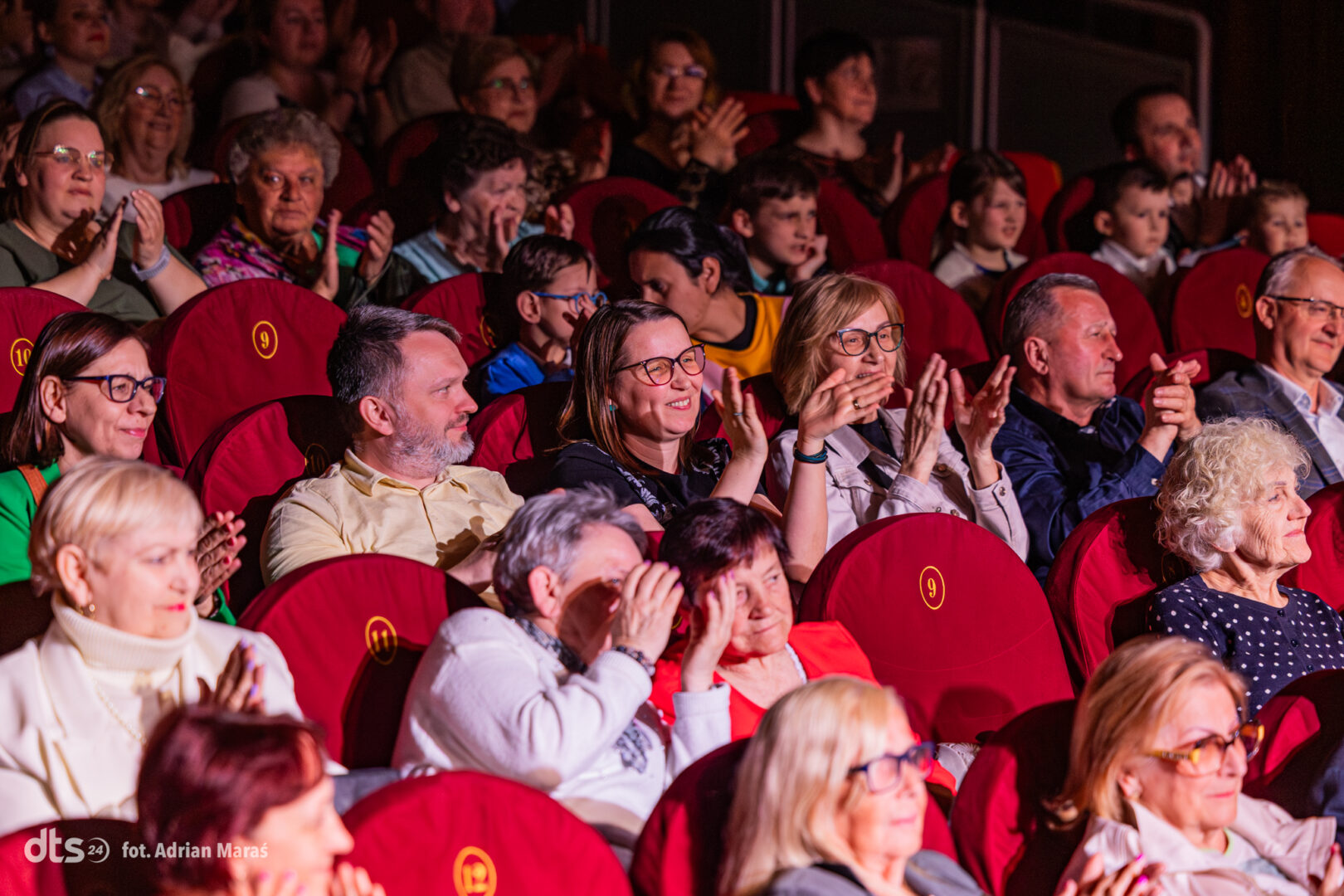 Audience in a theater, applauding and watching a performance from red seats.