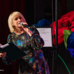 Female singer on stage, holding a microphone and a folder of sheet music, wearing a colorful floral dress against a dark backdrop with big red and blue fabric flowers nearby.