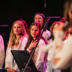 Group of young female singers in white blouses standing on a stage with microphones and a music stand under pink stage lighting.