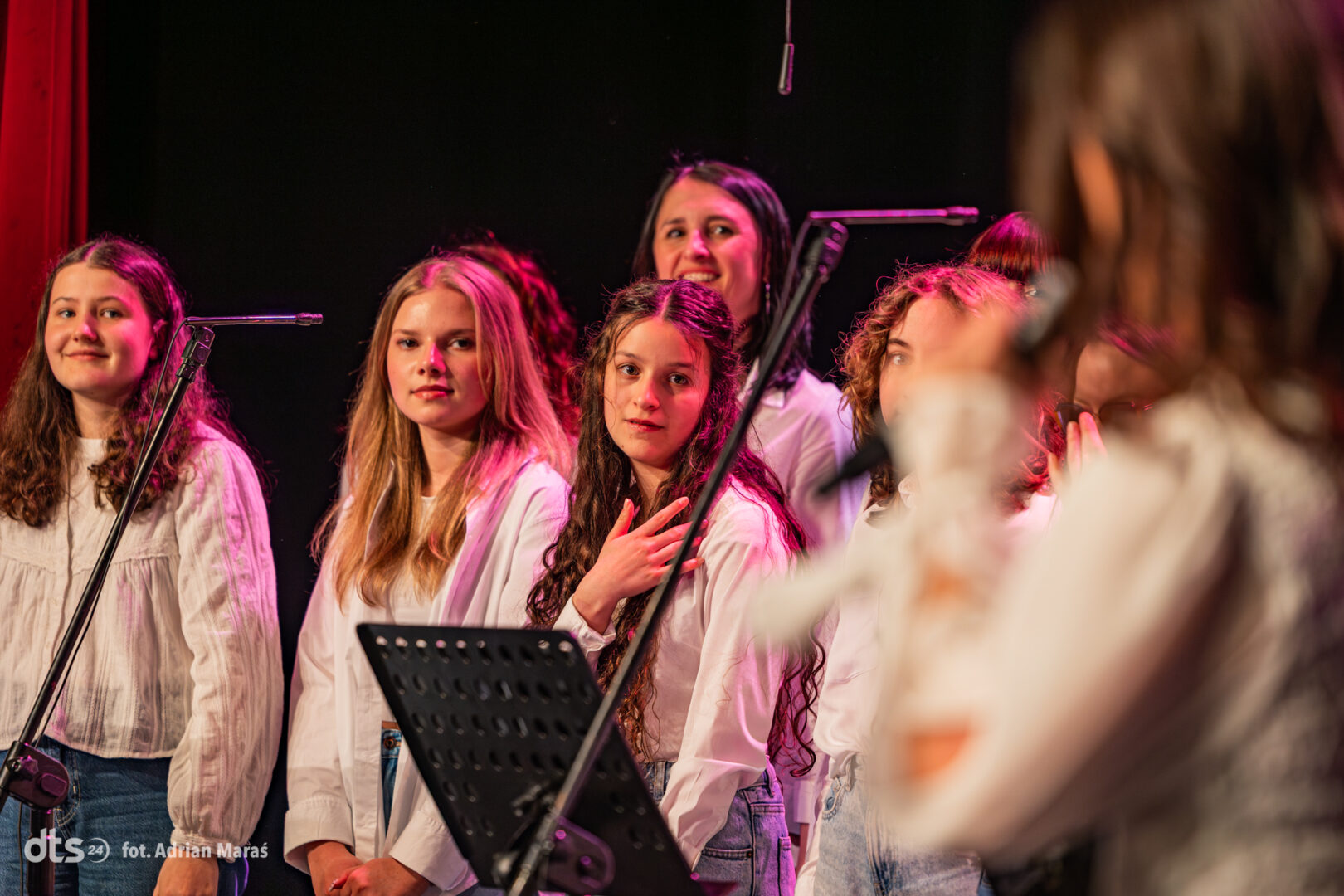 Group of young female singers in white blouses standing on a stage with microphones and a music stand under pink stage lighting.