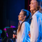 Two female singers on stage in white shirts, each holding a microphone, bathed in blue stage lighting.