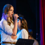 Female singer on stage holding a microphone, wearing a white shirt and jeans, bathed in blue stage lighting.