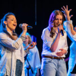 Group of female singers on stage, two in front holding wireless microphones, wearing white shirts and jeans.
