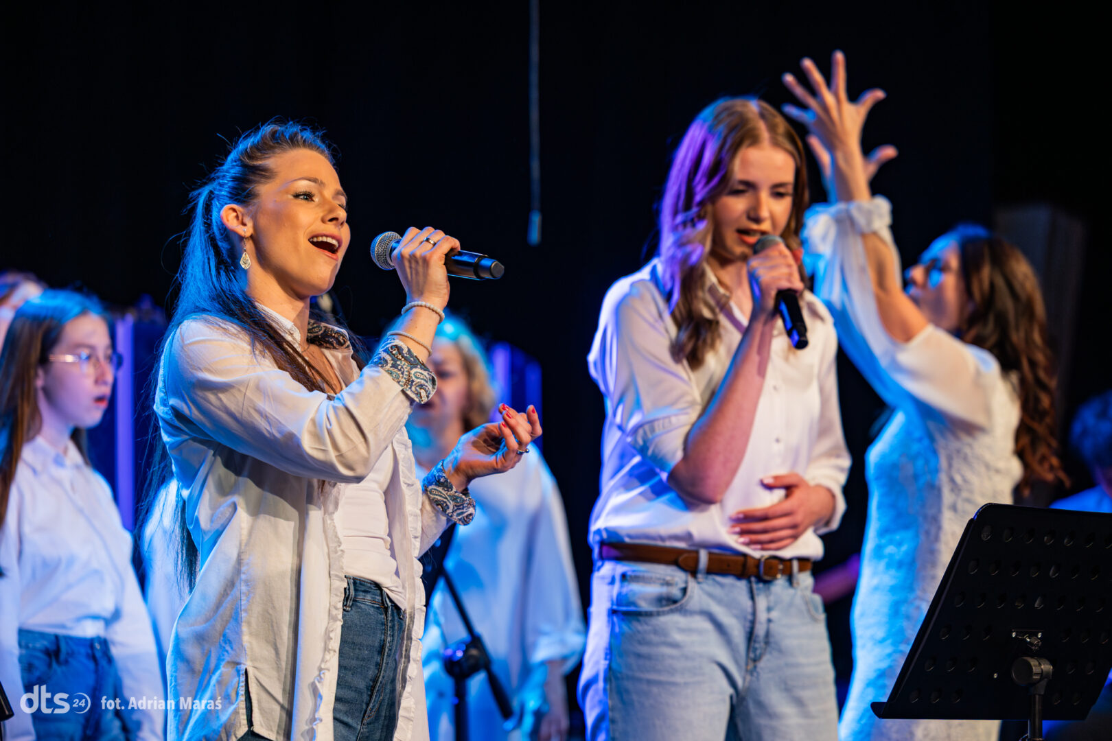 Group of female singers on stage, two in front holding wireless microphones, wearing white shirts and jeans.