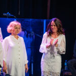 Group of female singers on stage in white outfits, with a guitarist in the background on the right.