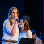 Young woman in a white shirt sings into a handheld microphone on stage, with teammates in white shirts smiling behind her.