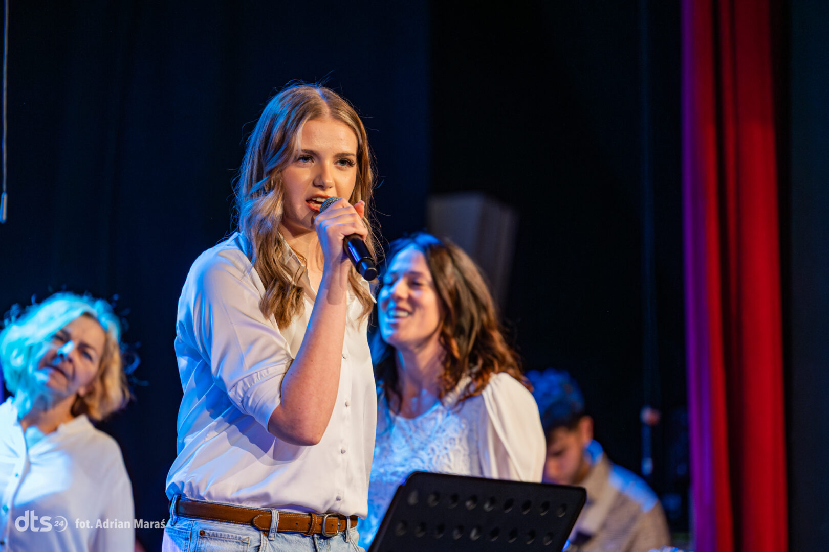 Young woman in a white shirt sings into a handheld microphone on stage, with teammates in white shirts smiling behind her.