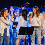 Two women sing into microphones at the front of a blue-lit choir on stage, with backup singers behind them.