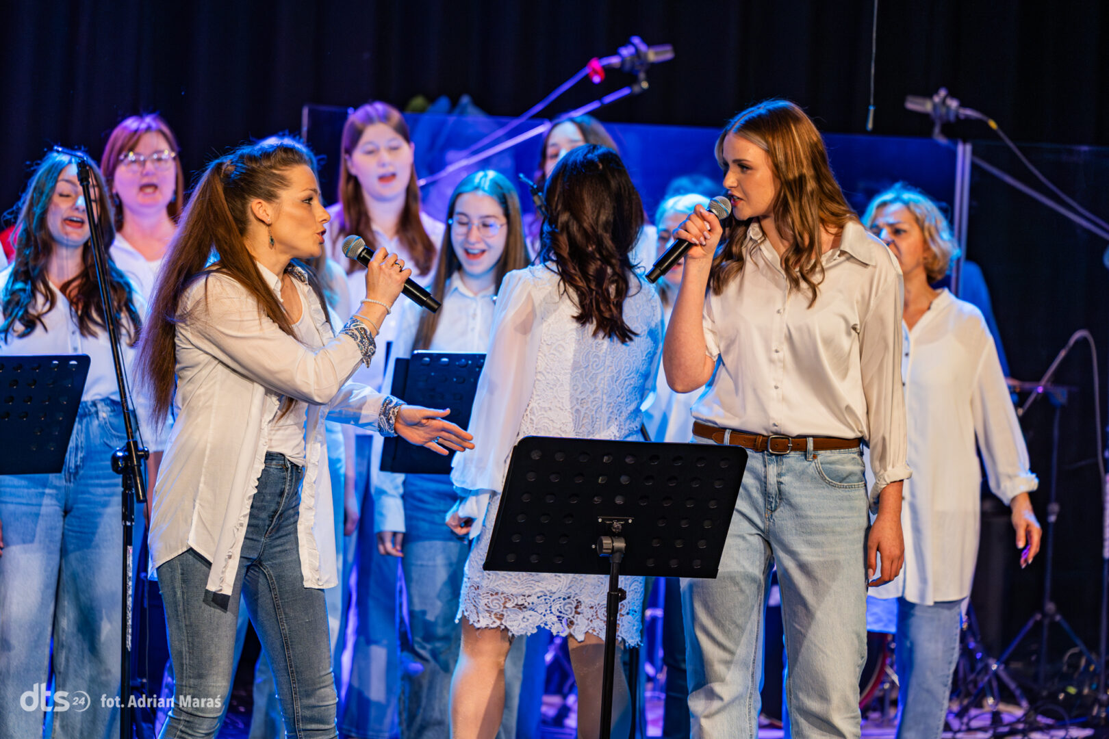 Two women sing into microphones at the front of a blue-lit choir on stage, with backup singers behind them.