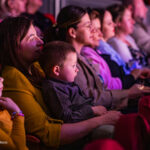 Audience watching a performance in a dimly lit theater, a mother in yellow holds a young child on her lap.