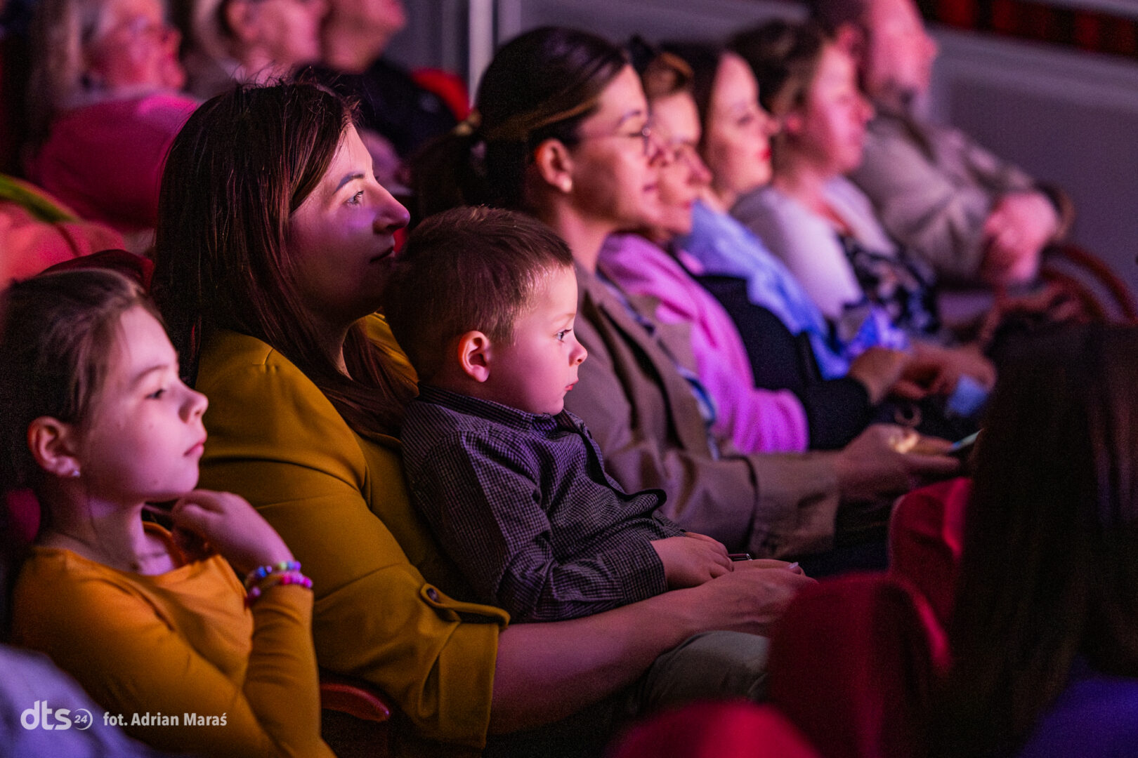 Audience watching a performance in a dimly lit theater, a mother in yellow holds a young child on her lap.