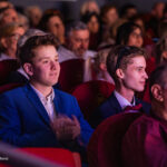 Two young men in formal attire sit in red theater seats, watching a performance among a crowded audience.