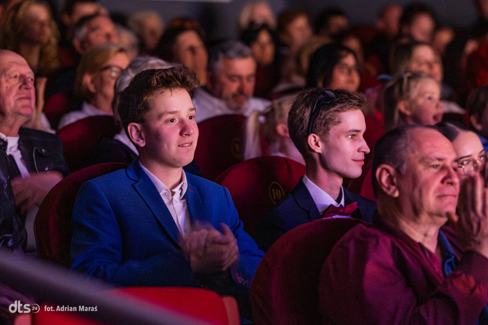 Two young men in formal attire sit in red theater seats, watching a performance among a crowded audience.