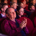 Middle-aged man in a maroon jacket sits in a theater, clapping as others watch a performance.