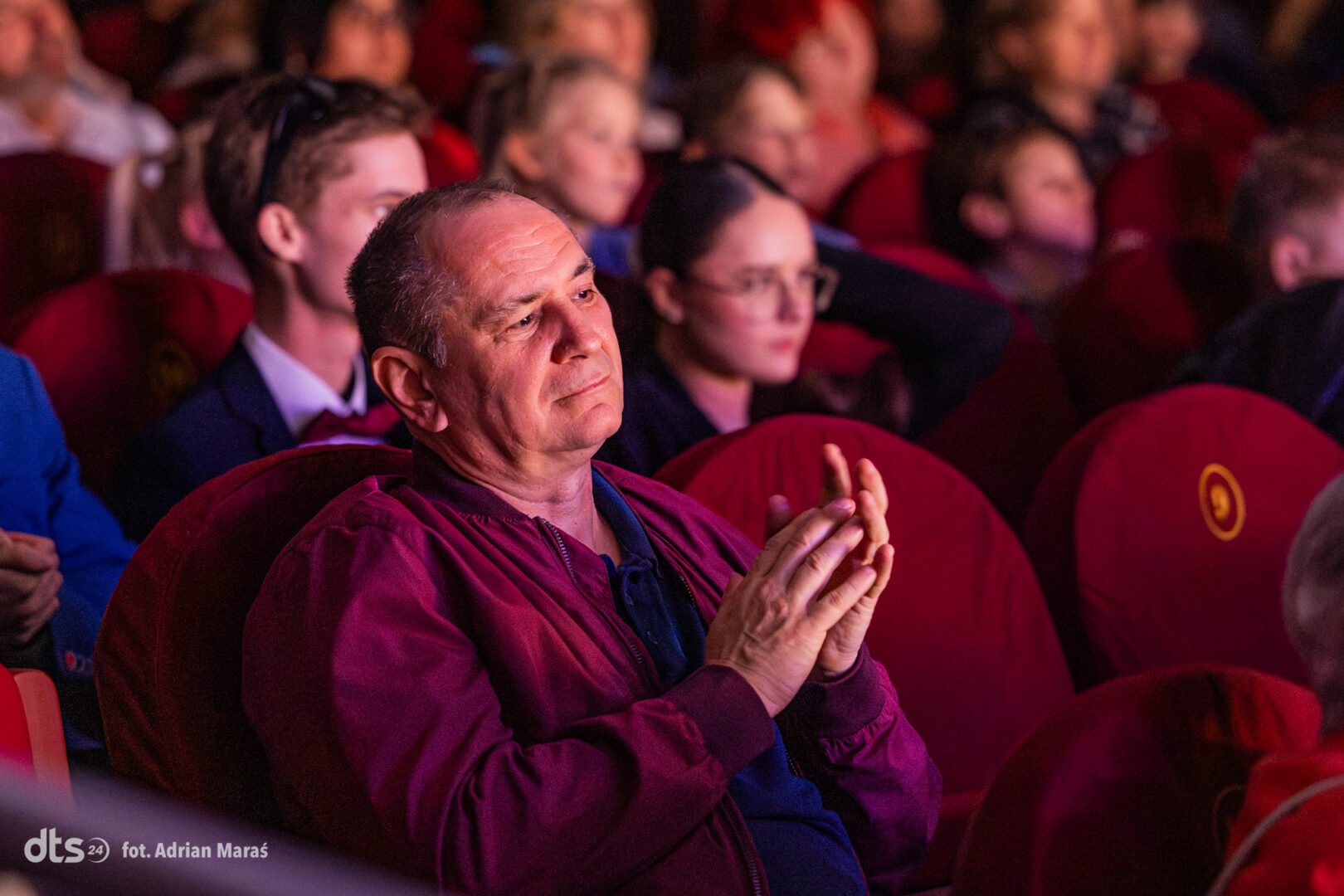 Middle-aged man in a maroon jacket sits in a theater, clapping as others watch a performance.