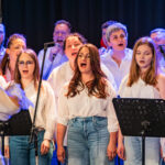 A choir of diverse performers wearing white shirts and jeans sings on stage, conductor visible on the left, microphones up front.