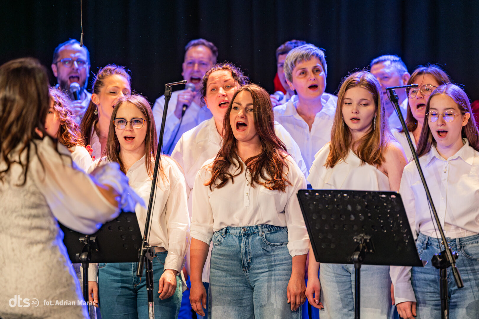 A choir of diverse performers wearing white shirts and jeans sings on stage, conductor visible on the left, microphones up front.