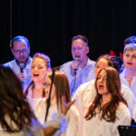 A choir of men and women in white shirts singing on a dimly lit stage, each holding a microphone.
