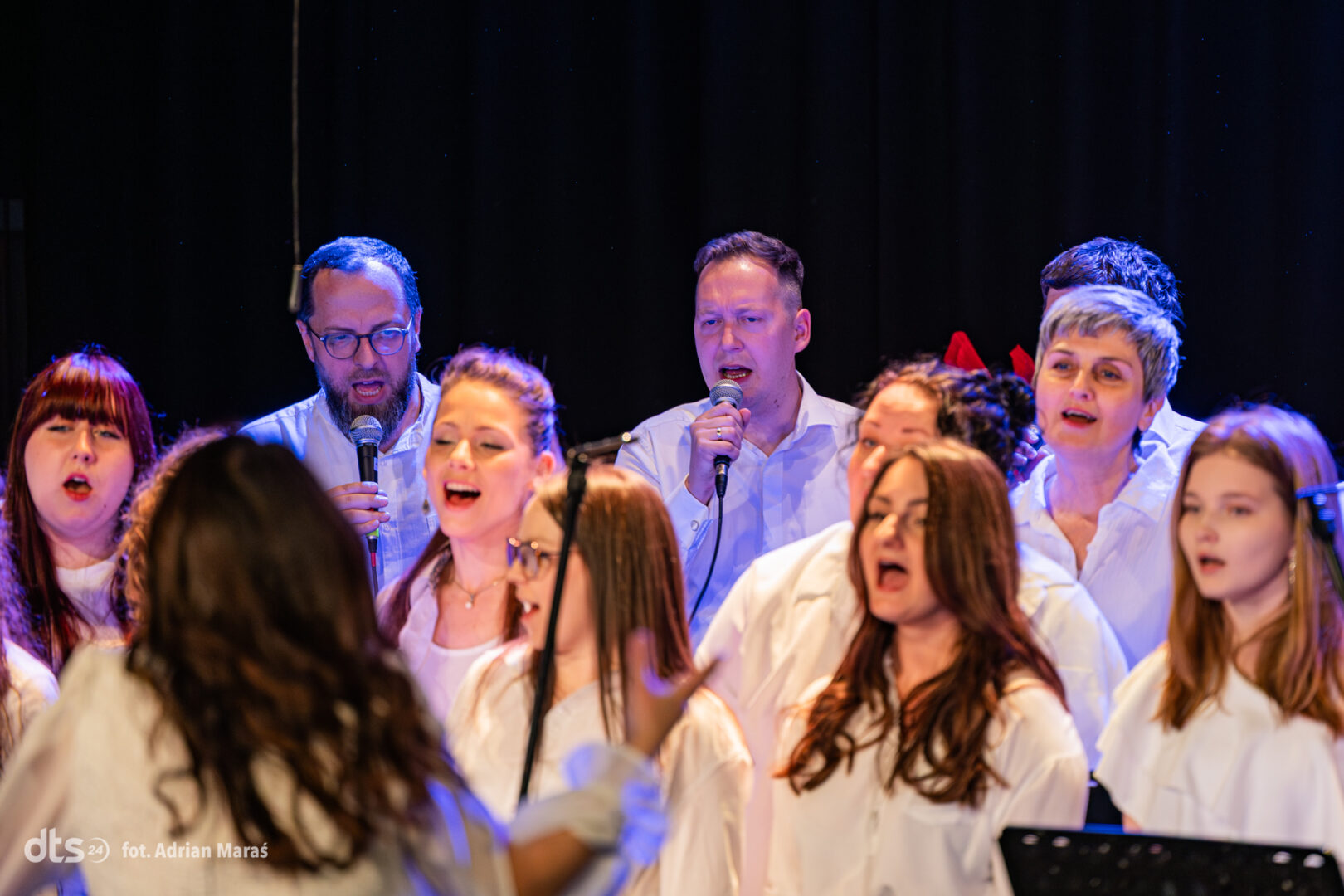 A choir of men and women in white shirts singing on a dimly lit stage, each holding a microphone.