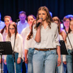 Lead female singer in a white blouse and jeans performing on stage with a choir behind her.