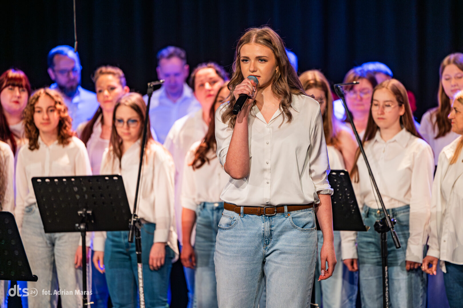 Lead female singer in a white blouse and jeans performing on stage with a choir behind her.