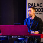 Male musician playing a red Nord Stage 4 keyboard on stage, wearing a black jacket and earphones, focused on performance.