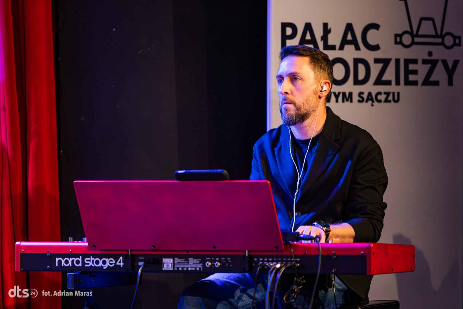 Male musician playing a red Nord Stage 4 keyboard on stage, wearing a black jacket and earphones, focused on performance.
