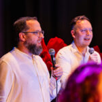 Two men in light shirts speak into handheld microphones on a dim stage with red flowers in the background? (informative alt)