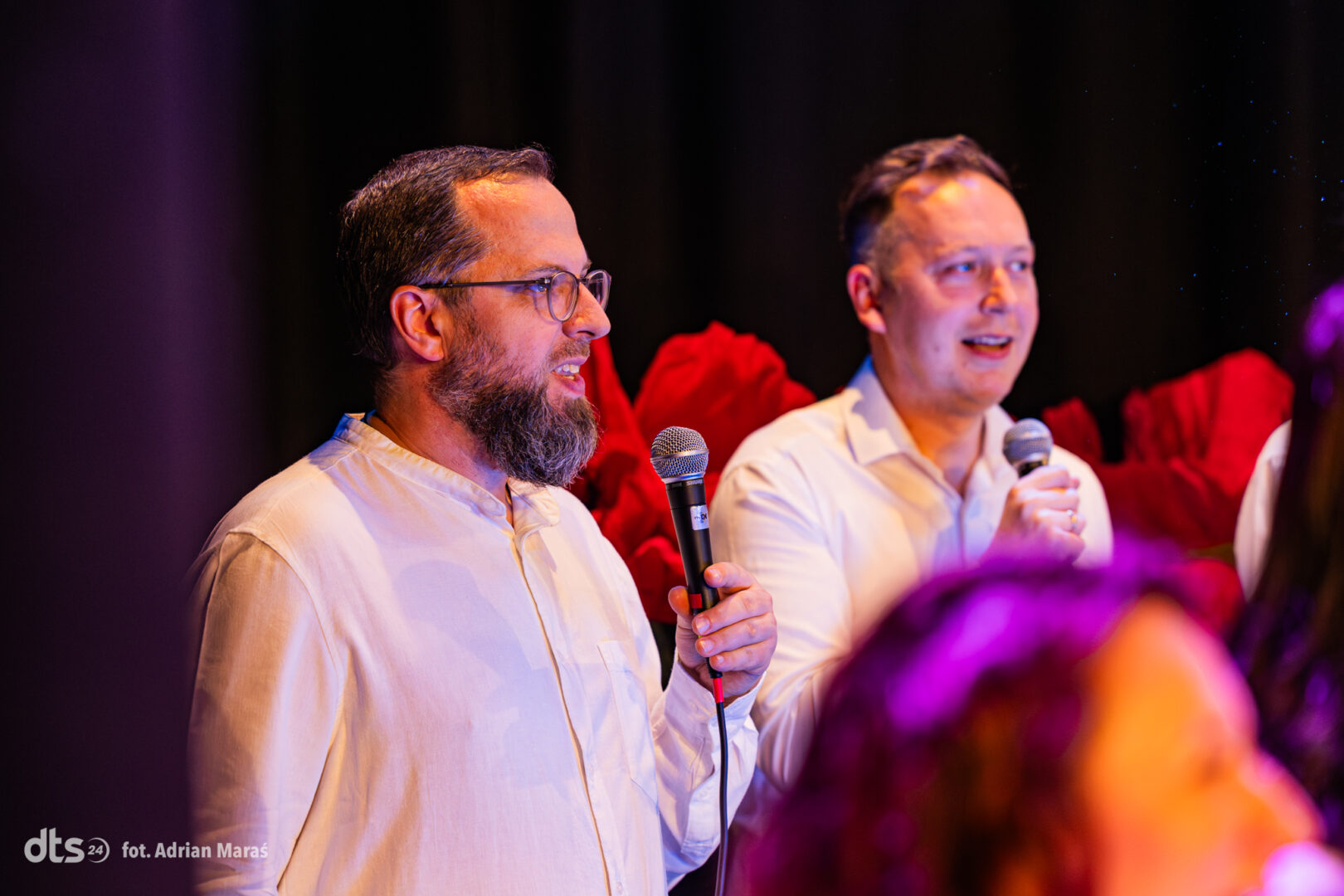 Two men in light shirts speak into handheld microphones on a dim stage with red flowers in the background? (informative alt)