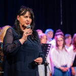 Woman in a dark blue blouse sings into a microphone on stage, with a choir in white behind her.