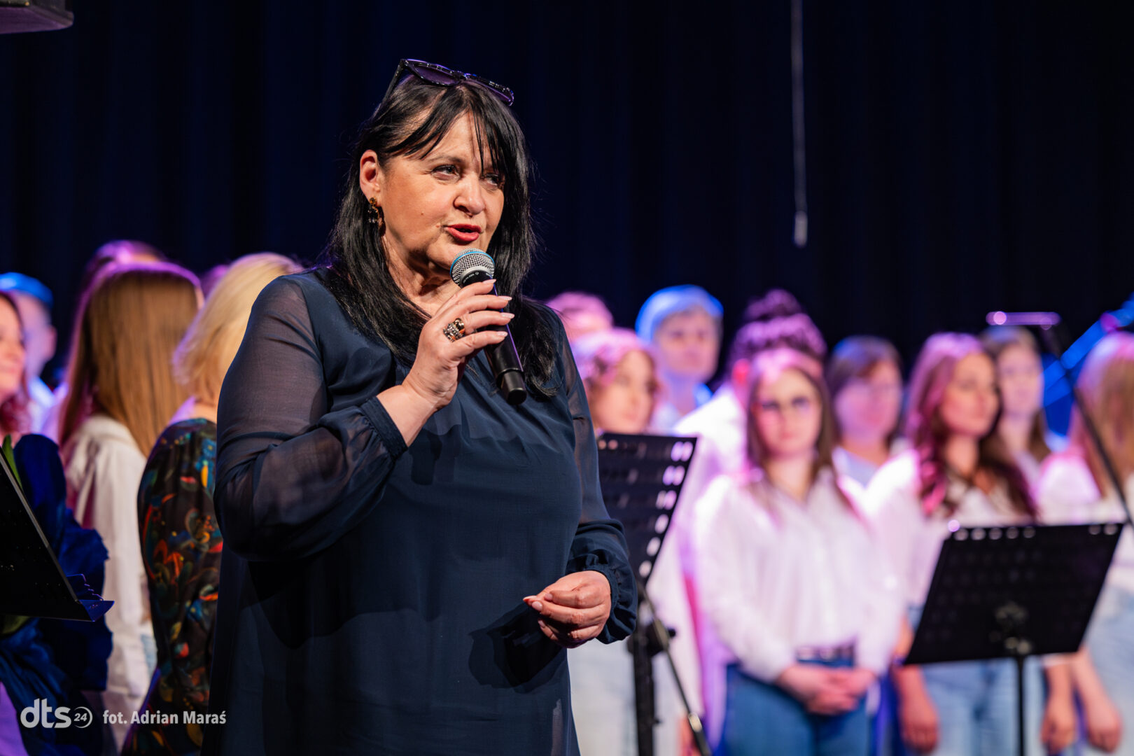 Woman in a dark blue blouse sings into a microphone on stage, with a choir in white behind her.