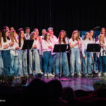 Young choir of girls and boys in white tops and jeans singing on stage with a female conductor nearby, microphones in front.