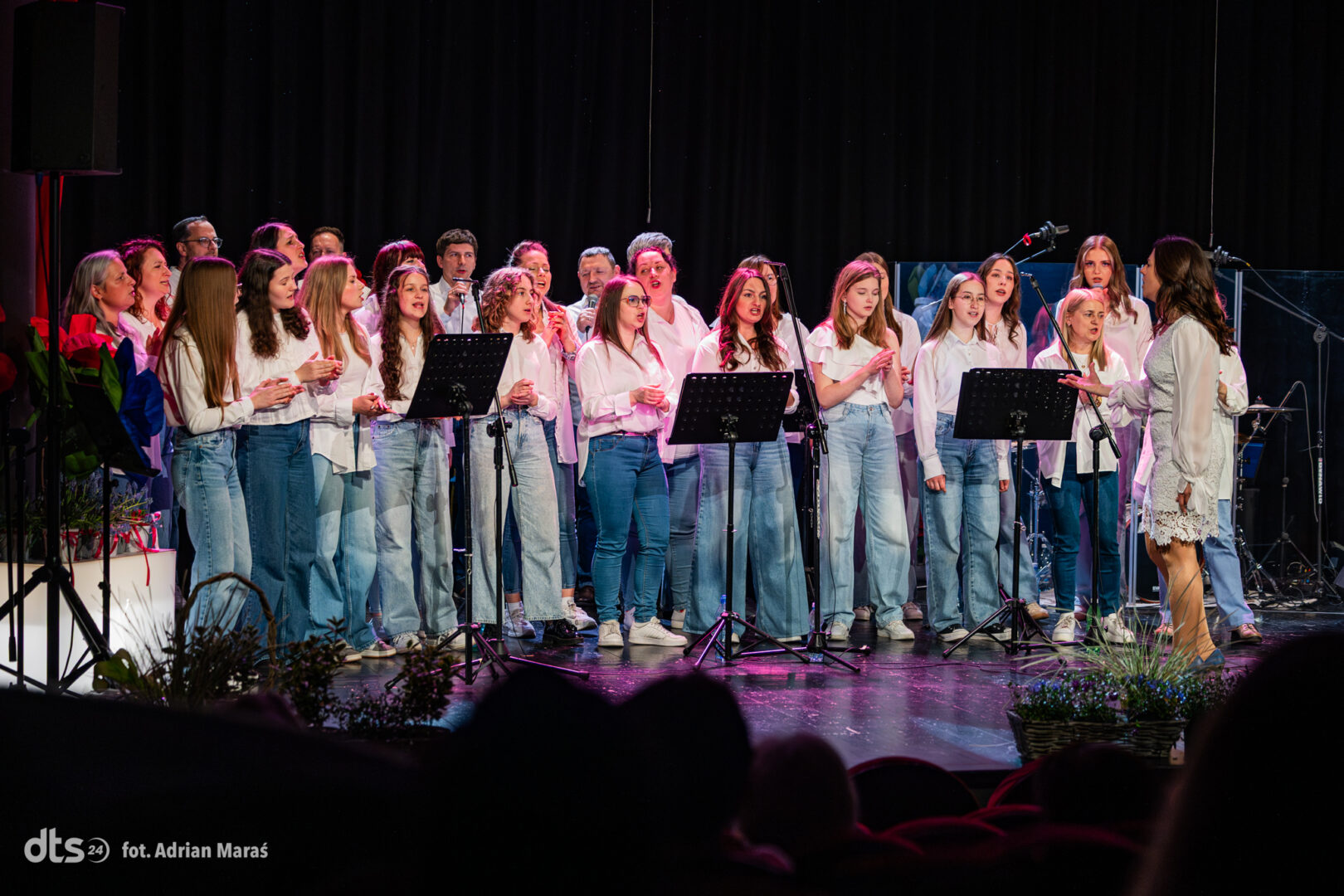 Young choir of girls and boys in white tops and jeans singing on stage with a female conductor nearby, microphones in front.