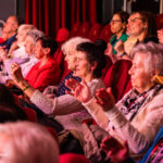 Audience in a theater clapping and cheering during a performance while seated in red seats with warm lighting.