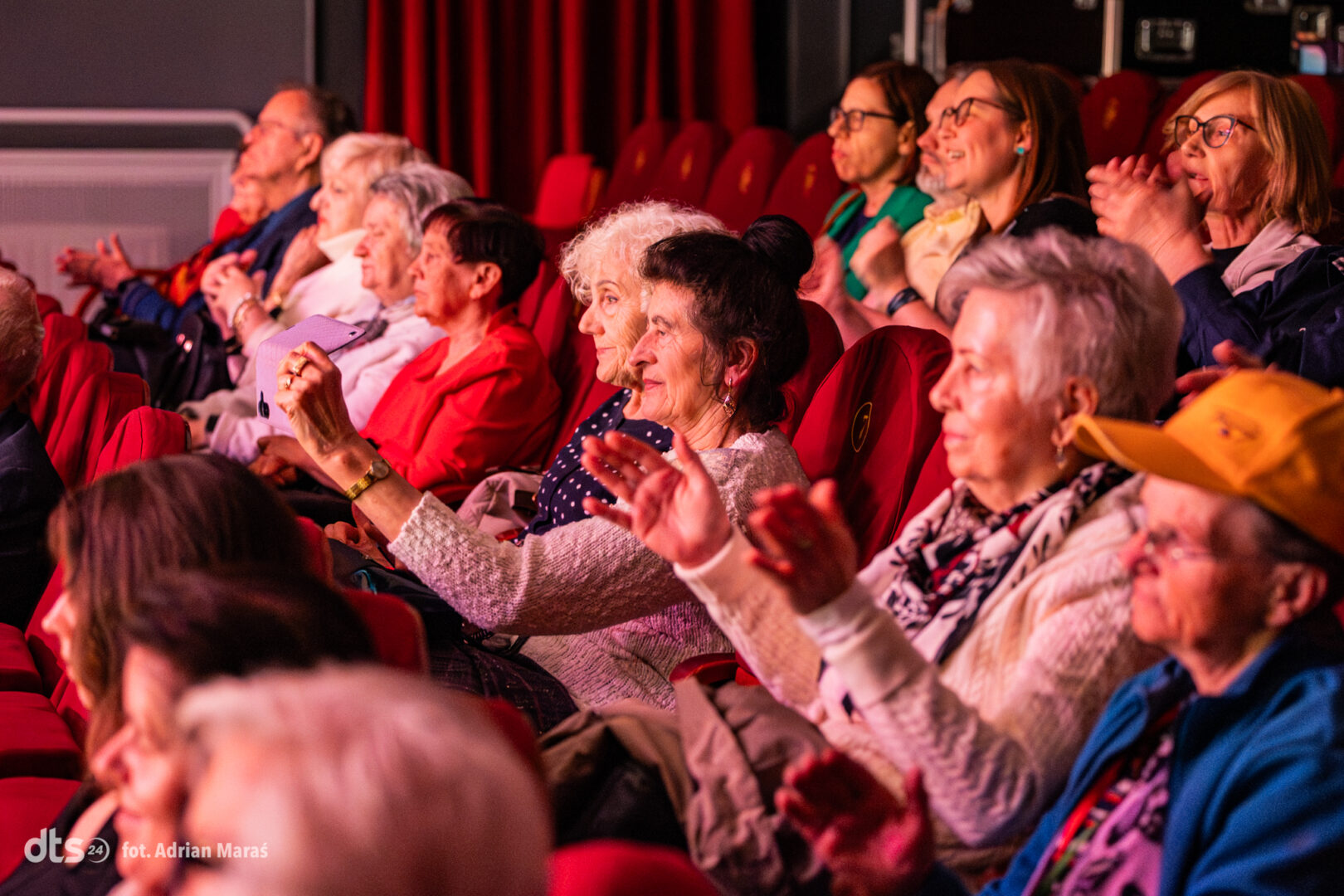 Audience in a theater clapping and cheering during a performance while seated in red seats with warm lighting.